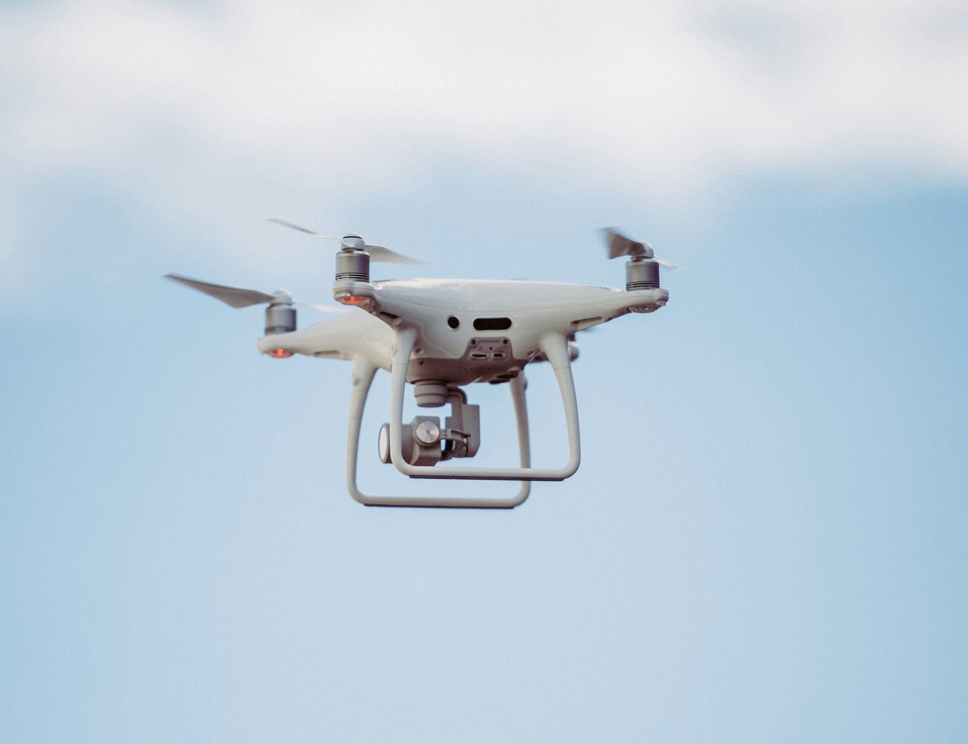 A white four-rotor camera drone hovering against a pale overcast sky, its gimbal camera pointed downward