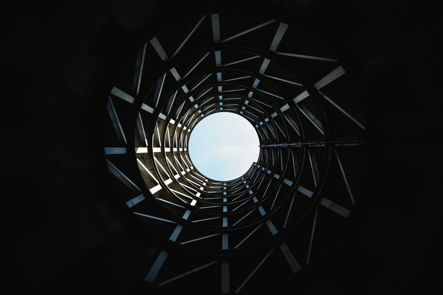 Looking straight up through the dark spiralling underside of a staircase toward a small circle of pale sky
