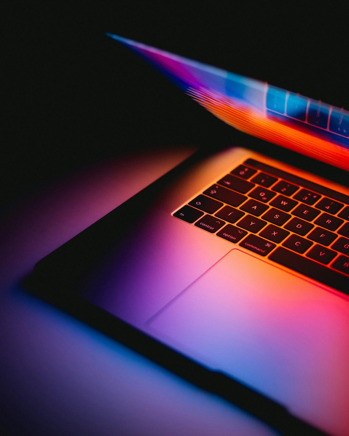 A half-closed laptop photographed in darkness, its keyboard and lid lit from below in orange, pink and blue