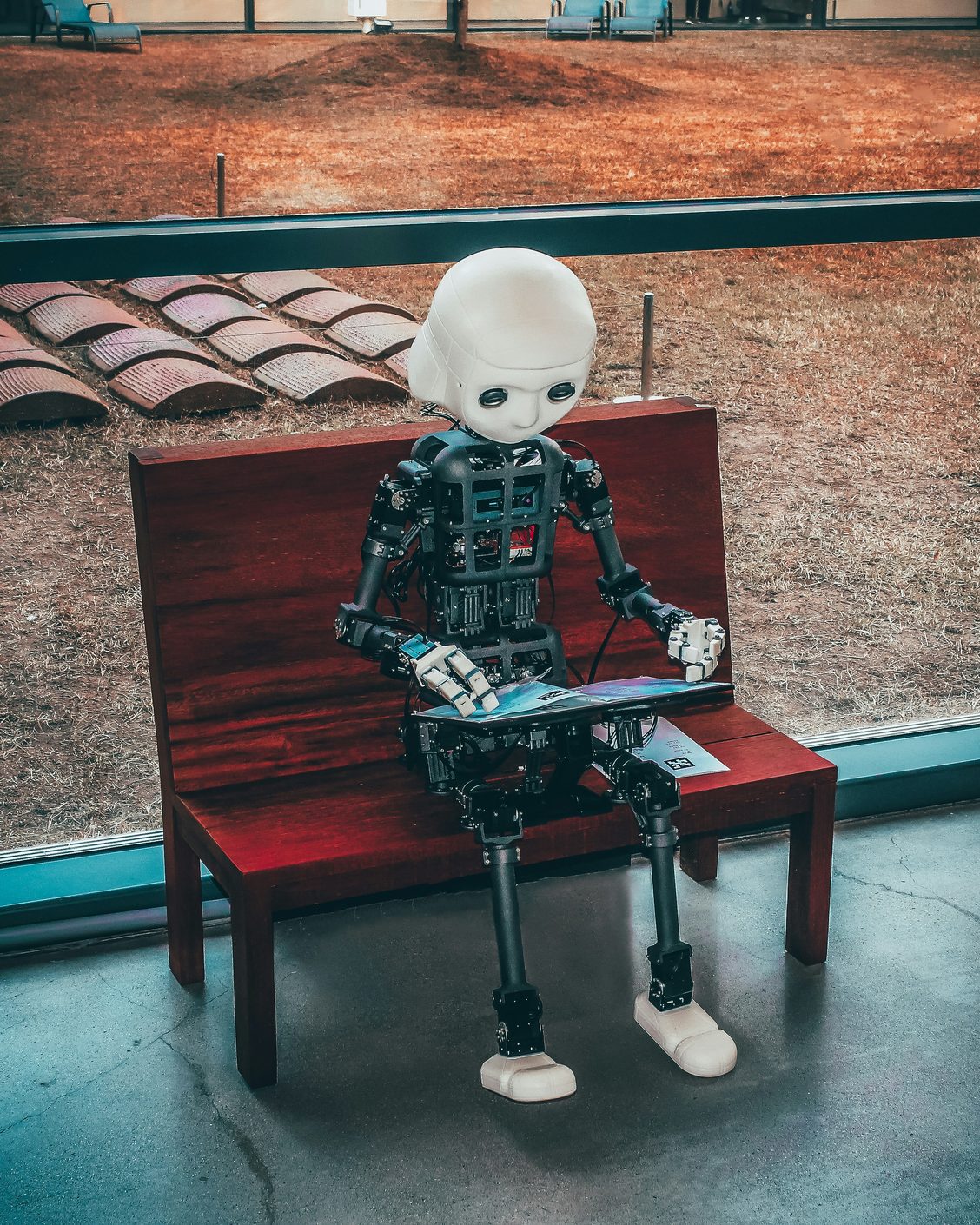 A small white and black humanoid robot sitting alone on a red wooden bench holding a tablet, seen through a window