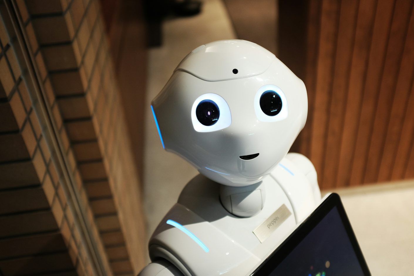 Close-up of a white humanoid robot with large round eyes standing indoors beside a brick wall
