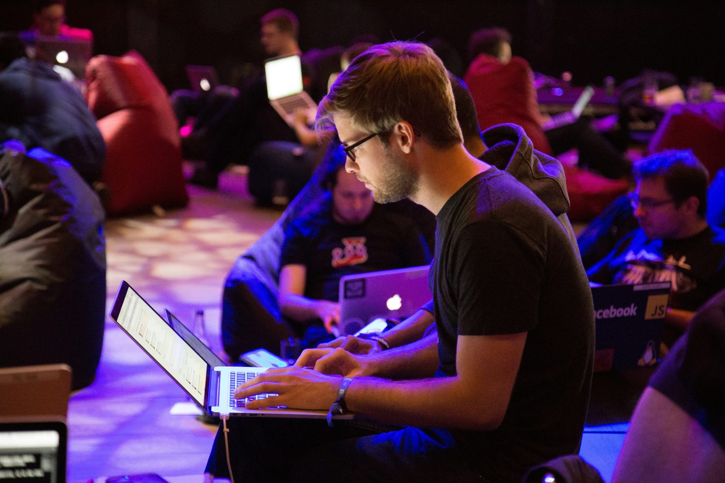 A man in glasses typing on a laptop among seated people in a dimly purple-lit room