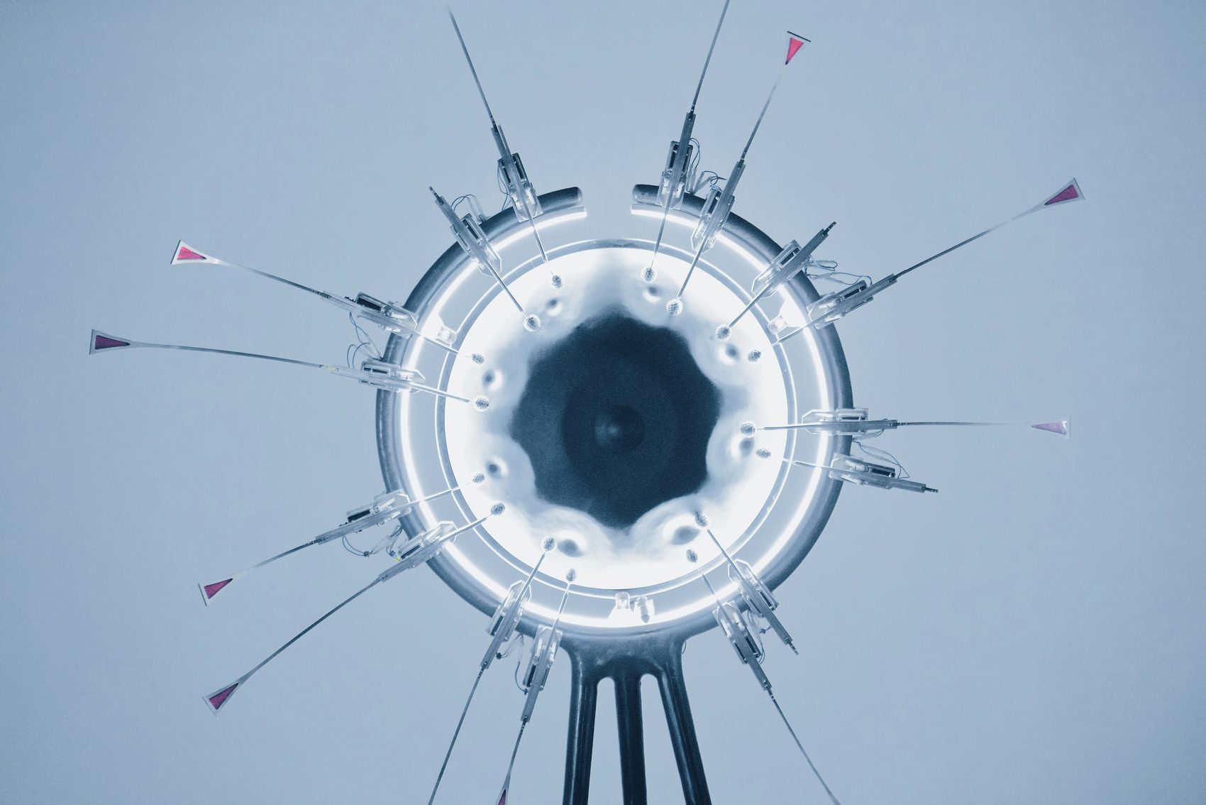 Overhead view of a glowing ring light with thin instruments radiating outward on a pale blue background